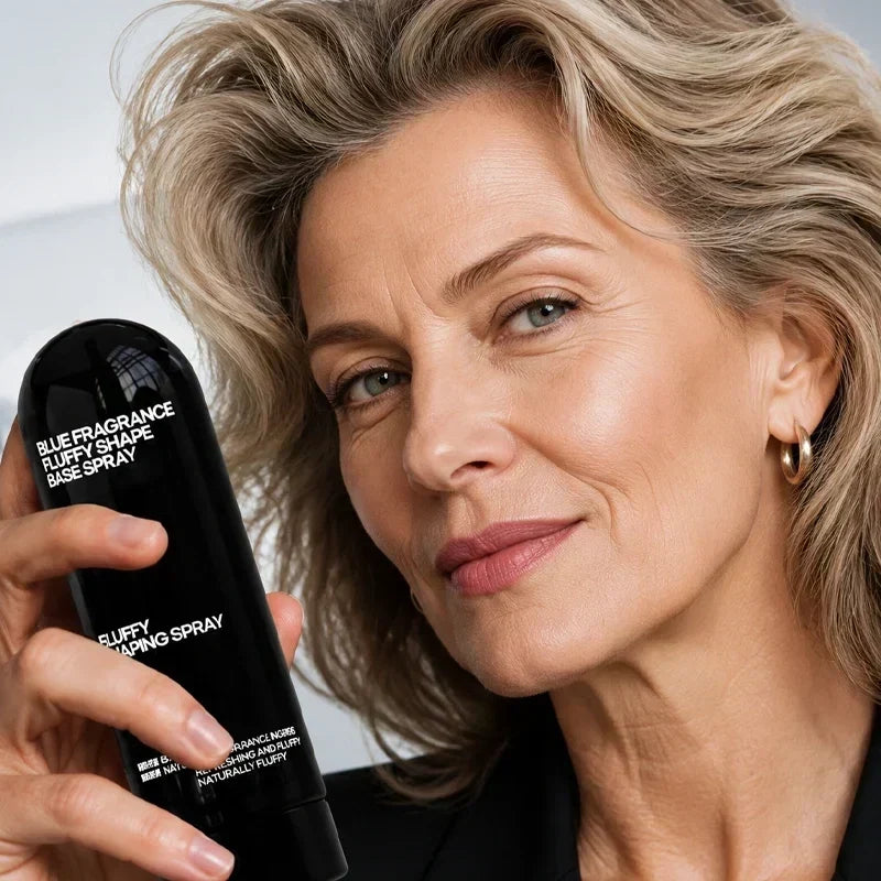 Silver-haired woman gently lifting her hair in front of a mirror with the styling spray on the counter